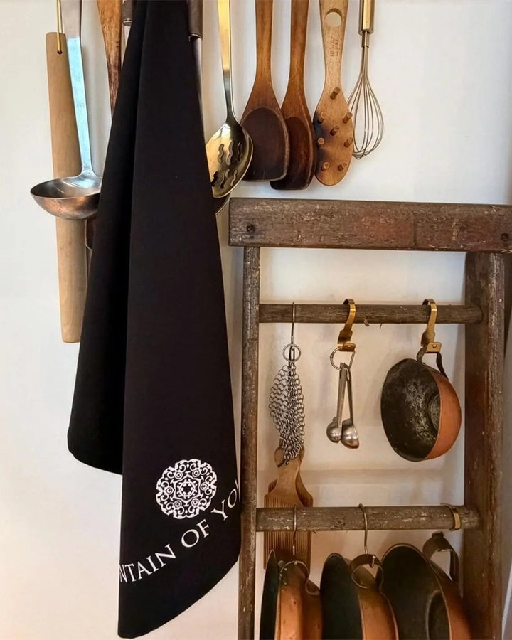 The black FoYL tea towel hanging in a kitchen beside copper pans